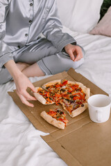 Female hand holding a slice of pizza Model sits on the bed in gray pajamas