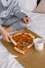 Female hand holding a slice of pizza Model sits on the bed in gray pajamas