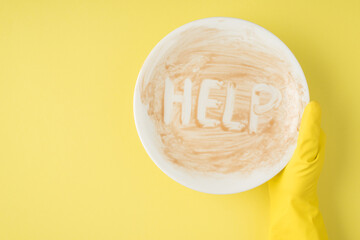 Top view photo of hand in yellow rubber glove holding dirty dish with inscription help on isolated yellow background with copyspace