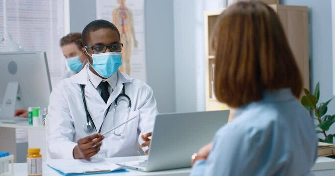 Young African-American doctor in protective medical face mask sits in office and listens carefully to patient talks about symptoms that bother her.