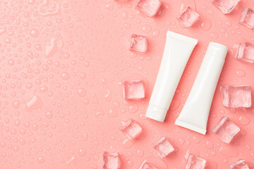 Top view photo of two white cream bottles without label ice cubes on the right and drops on isolated pastel pink background with blank space on the left
