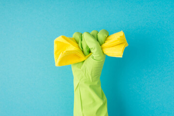 Photo of raised hand in green glove clenching yellow rag into fist on isolated blue background with copyspace
