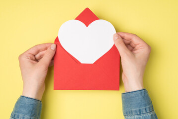 First person top view photo of woman's hands holding open red envelope with white heart on isolated...