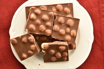 Several slices of dark chocolate with nuts on a white plate on a red linen napkin, close-up, top view.