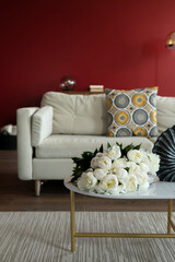 bouquets of white peonies lie on the coffee table. In the background is a white sofa with a colorful pillow. The living room has red walls. A bouquet of white flowers. Fresh flowers in the interior.