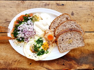 Pyszne śniadanie, jajka smażone, sałatka, serek i ciemne pieczywo na białym talerzu na tle drewnianego stołu.
