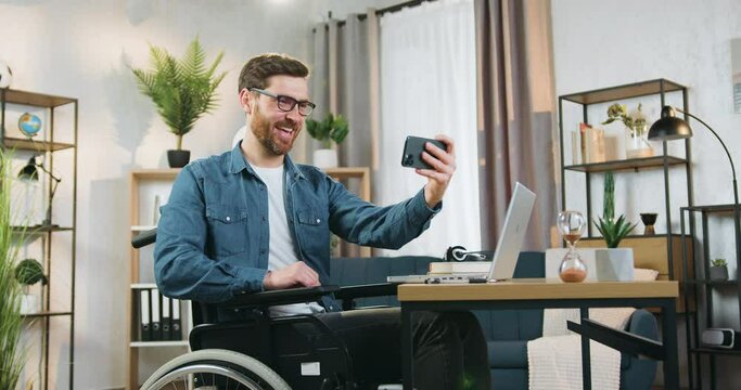 Attractive cheerful carefree adult bearded handicapped man in glasses sitting in wheelchair after accident and making selfie on smartphone at home