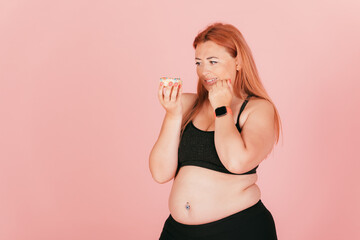 Oversized young woman wearing sports outfit looks at sweet donut junk food