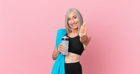 middle age white hair woman feeling proud,smiling positively with thumbs up with a towel and water bottle. fitness concept