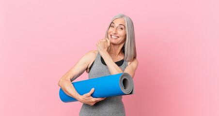 middle age white hair woman feeling happy and facing a challenge or celebrating and holding a yoga mat. fitness concept