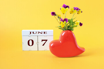 Calendar for June 7 : the name of the month of June in English, the numbers 0 and 7, a vase in the shape of a red heart with a bouquet of violets on a yellow background