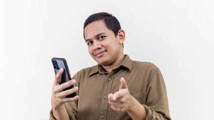 Young Asian Malay man smiling while holding a smartphone and pointing at the camera.