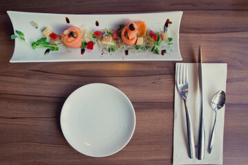 Smoked salmon on a long, white plate with an empty plate, fork, knife, spoon and napkin on a wooden table.
