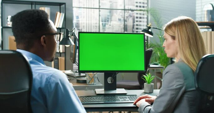 Two multiracial workers sitting at office and discussing business issues together. Blonde woman pen and typing at the computer green screen at the workplace. People looking at each other
