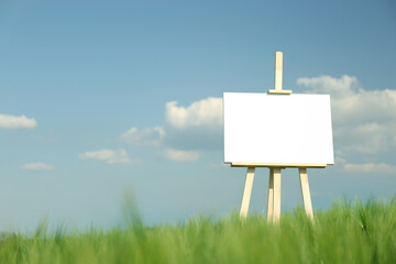 Wooden easel with blank canvas in picturesque green field on sunny day. Space for text