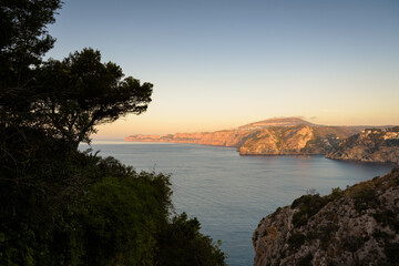 Sunrise light illuminates the sea and rocky cliffs of the Mediterranean coast on a summer day, Javea, Spain