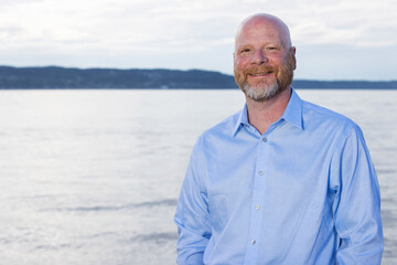 Environment portrait of bald man with a beard outside in front of ocean water. Business casual head shot wearing a button up shirt with open collar.  - Powered by Adobe