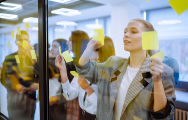 Business colleagues writing on sticky note on glass wall with coworkers standing by in office....