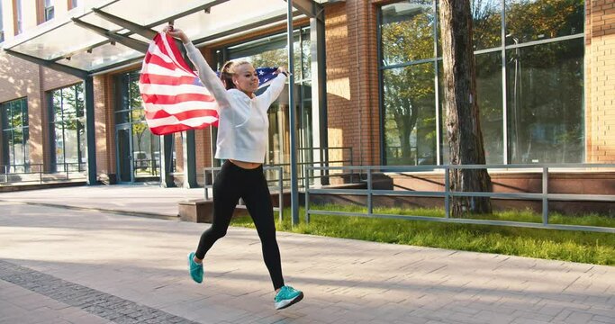 Cheerful Caucasian young pretty woman runner jogging outdoors in town and smiling holding American flag above. Happy beautiful fit female with USA flag running outside. Sunny day, patriotism concept