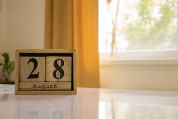 Wooden blocks of the calendar represents the date 28 and the month of August on the background of a window, curtain and a plant.
