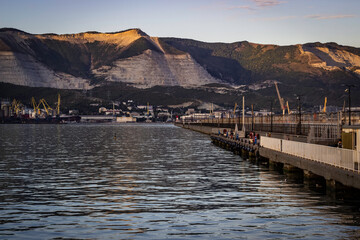 The anti-wave breakwater of the seaport with a lighthouse extending to the center of the frame with the mountains and the port in the background.