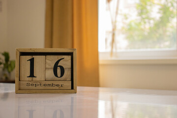 Wooden blocks of the calendar represents the date 16 and the month of September on the background of a window, curtain and a plant.