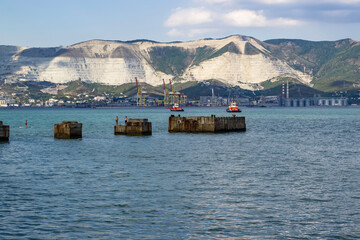 Debris of the old pier leaving in the sea bay. Mountains, port and tugboat in the background.