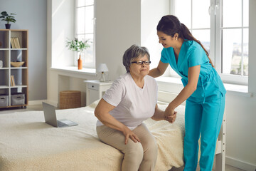 Smiling caring nurse supporting patient assisting senior woman to get up from bed helping elderly...
