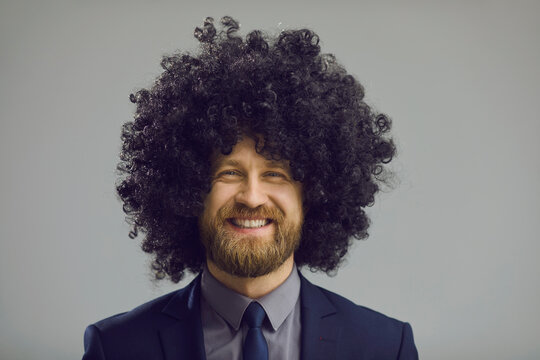 Headshot of cheerful man with crazy hair smiling at camera. Studio portrait of happy positive bearded young businessman in suit, necktie and funny curly Afro style wig