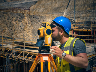 Land surveyor stands by theodolite on a tripod against construction site at background. Topographer...