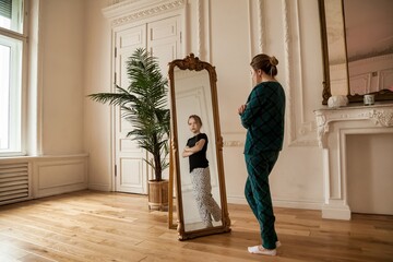 Middle age woman looking at mirror in living room and sees her daughter in reflection - obrazy, fototapety, plakaty