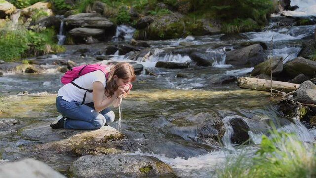 Woman drinking fresh water from mountain river stream. Traveler drinks clean water from forest river