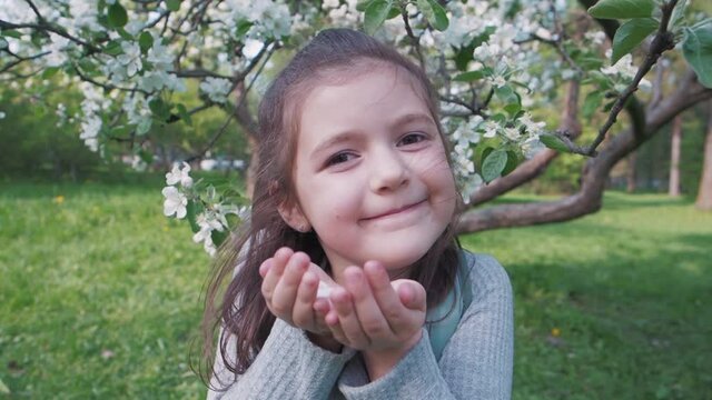 Close-up. Cute brunette little female kid blows off petals of white apple tree flowers, smiles and looks at the camera. Happy childhood in the flowered garden during spring time enjoy fresh nature