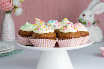 Carrot Cake Cupcakes on a cake plate with an Easter Bunny and a vase of flowers in the background.