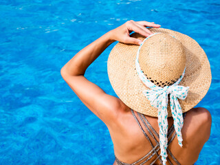 Woman in bikini with a hat from the back sitting on the edge of the pool.