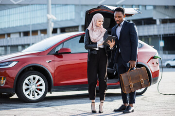 Muslim woman in hijab using digital tablet while talking with african businessman on suit. Female seller and male client standing near electric car and discussing all details of expensive purchase.