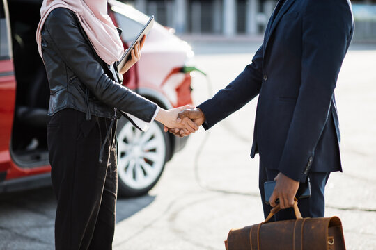 African businessman shaking hands with muslim woman in hijab after successful deal. Red electric car charging on background. Concept of people, selling and purchase. - Powered by Adobe