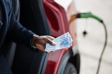 Close up of male hand holding money cash over blur background of charging electric car. Smart...