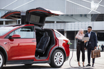Red electric car is charging while muslim woman and african man talking near. Female seller in hijab using laptop while demonstrating modern auto to male customer in suit.