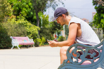 Fototapeta premium Young man holding cellphone sitting on park bench on sunny day. Boy on sleeveless shirt and shorts using smart phone outdoors in summer time