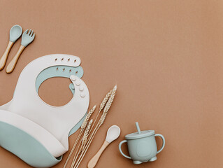 Composition with baby food accessory. Silicone cute dishware, bib and bowl on brown background. Neutral pastel colors