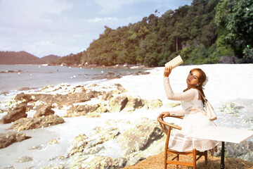 A beautiful Asian woman sitting on a table at a rock and holding a book and stretching it to block sunlight.