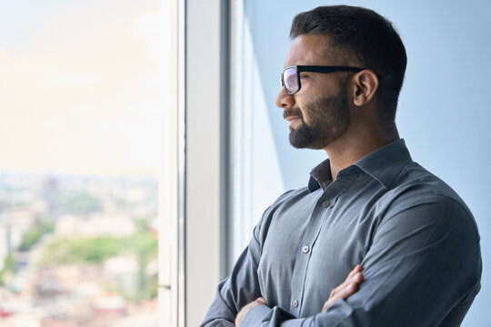 Headshot portrait of indian confident successful businessman in glasses thinking imagining future corporate financial career standing at office looking at window. Business concept.