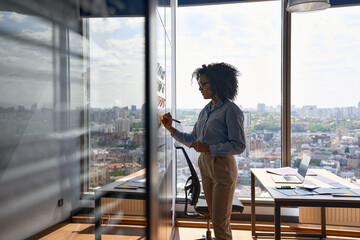 Young African american executive manager businesswoman writing strategy ideas on sticky notes on whiteboard doing business project presentation in contemporary corporate office with panoramic view. - Powered by Adobe