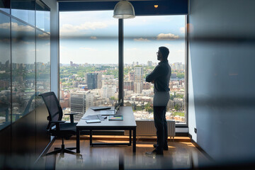 Silhouette of indian confident successful businessman thinking imagining future corporate financial career standing at office near window of high floor building with city view on big urban center.