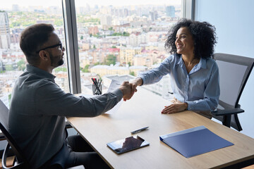 Indian friendly ceo businessman hr director holding job contract hiring welcoming female African American applicant manager shaking hands in contemporary office. Human resources concept.