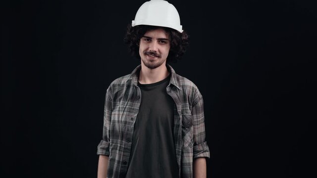 The charismatic hipster with a white engineer's helmet on his head flirts with eyebrows and looks forward proudly. Isolated on black background. The concept of life. People's emotions. 4k portrait