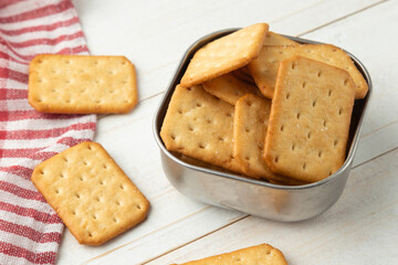 Cracker cookies in a stainless steel bowl with tablecloth on white wooden table background.