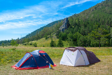Two open tents stand on grass in the mountains. Big rock and blue sky on background. Hiking camp.