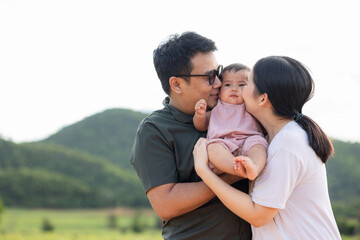 Happy asian family father mother child daughter on nature background . Parents embracing kissing little baby in arm standing outdoor with sunny day.